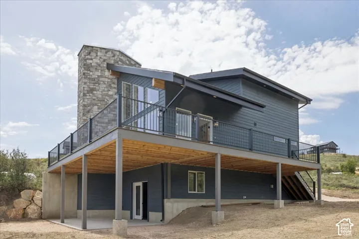 Modern exterior featuring dark siding, a stacked stone chimney, an expansive elevated deck with metal railings, and a covered patio beneath