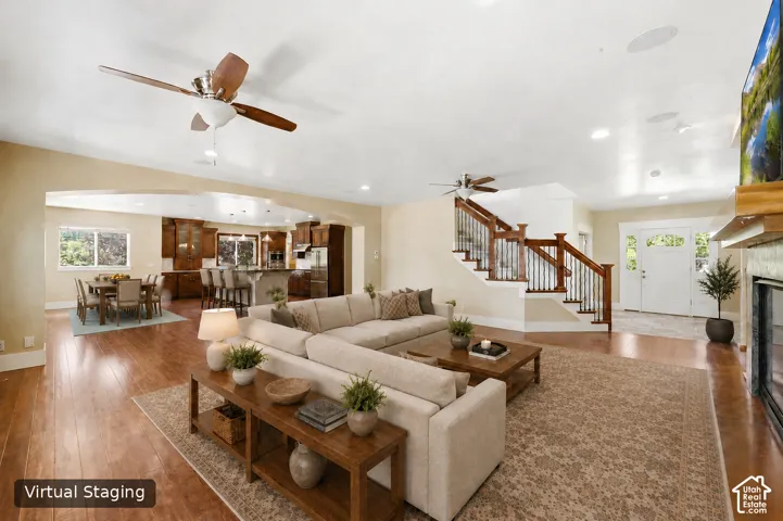 Expansive open-concept living area featuring wood-finish flooring, a traditional fireplace, a grand staircase with dark wood railings, a ceiling fan, and recessed lighting