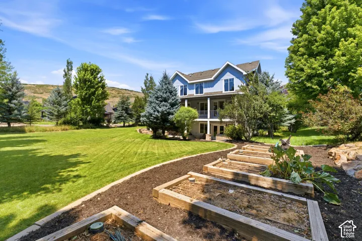 Expansive lawn area with a winding mulched path, tiered raised garden beds, mature trees, and a two-story home with a lower-level patio