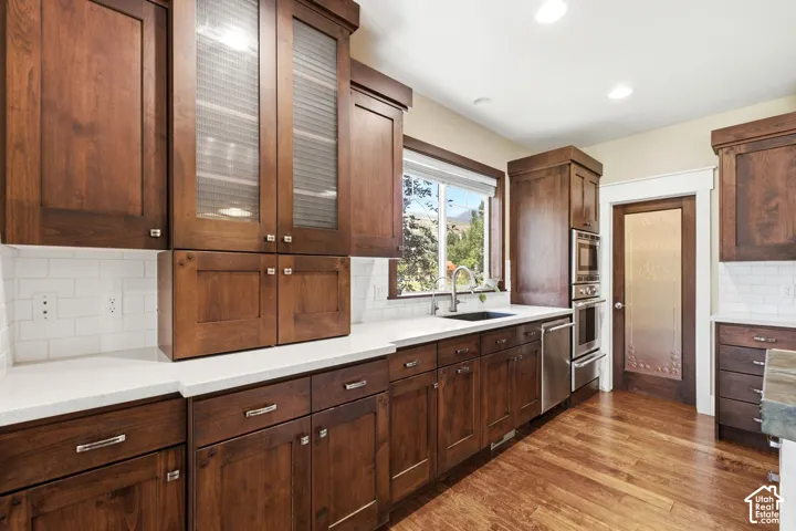 Wood-finish flooring, dark wood cabinetry, white subway tile backsplash, stainless steel double oven, and a built-in sink