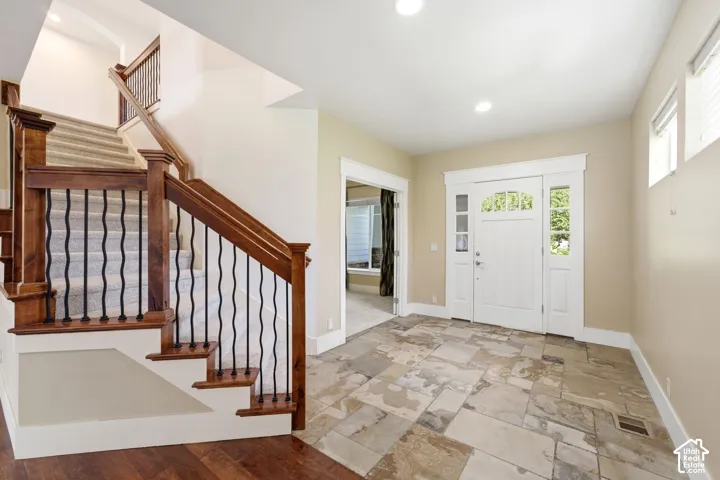 Two-story staircase with wood newel posts and wrought iron balusters, natural stone tile flooring, recessed lighting, and a white paneled entry door with sidelights