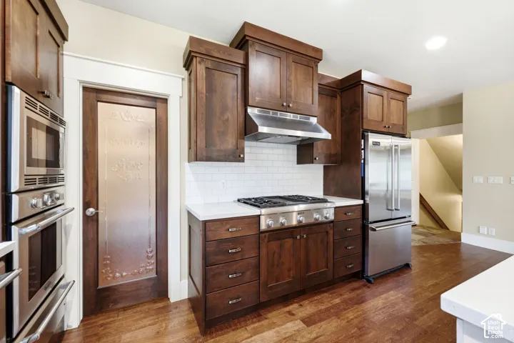 Kitchen featuring wood-finish flooring, dark wood cabinetry, white subway tile backsplash, and stainless steel appliances including a built-in oven and microwave