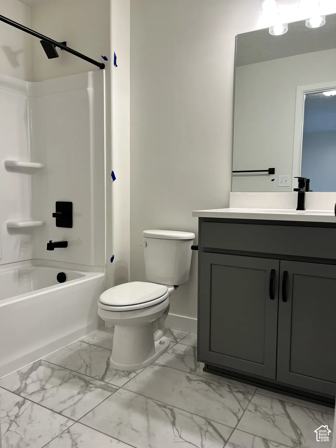 White bathtub and shower combination with matte black fixtures, a white toilet, gray vanity with black hardware, white countertop, and white tile flooring with gray veining