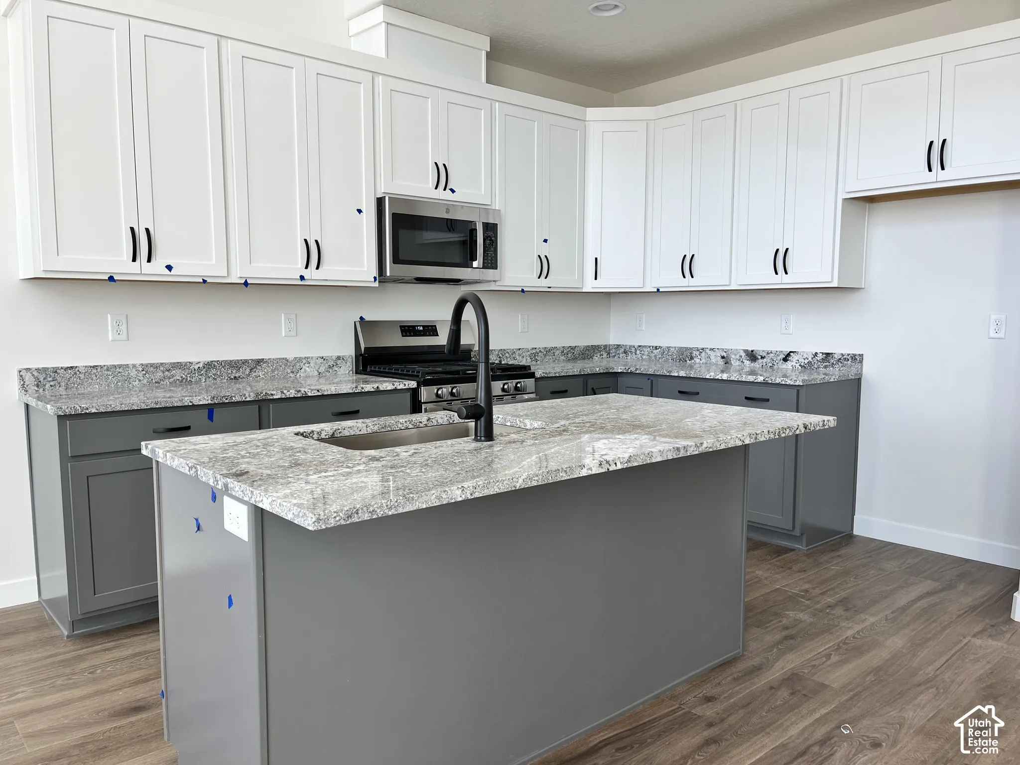 Two-tone cabinetry with white upper and gray lower cabinets, a kitchen island with an integrated sink, stainless steel appliances, wood-finish flooring, and light-toned countertops