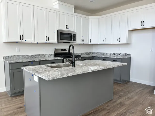 Two-tone cabinetry with white upper and gray lower cabinets, a kitchen island with an integrated sink, stainless steel appliances, wood-finish flooring, and light-toned countertops