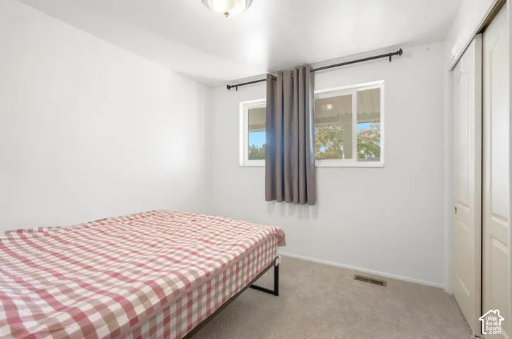 White walls with gray carpeting, featuring a double window with blinds and gray curtains, a flush-mount ceiling light, and a built-in closet with bi-fold doors