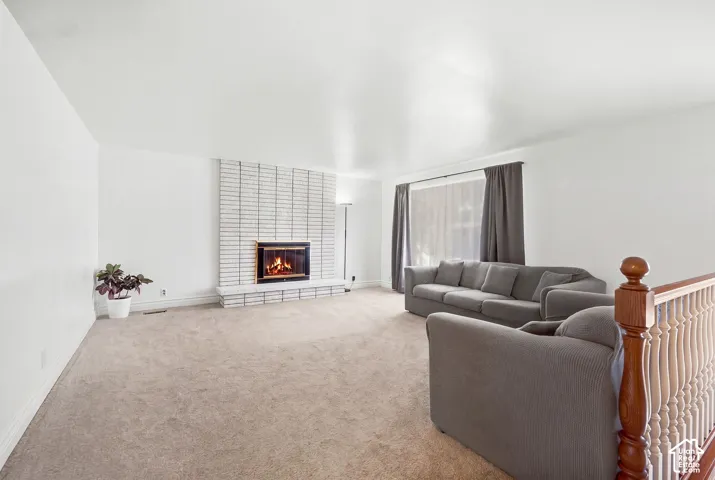 Carpeted living space featuring a white brick fireplace with a firebox, a large window, and a wooden banister