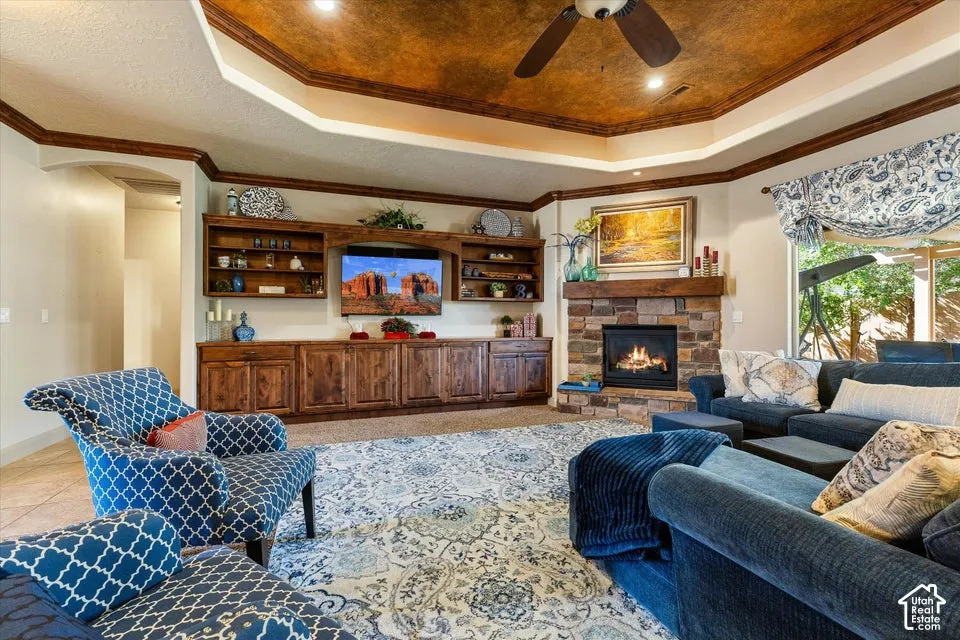 Tray ceiling with wood-finish trim, stone-clad fireplace, built-in wood cabinetry, integrated ceiling fan, and large windows