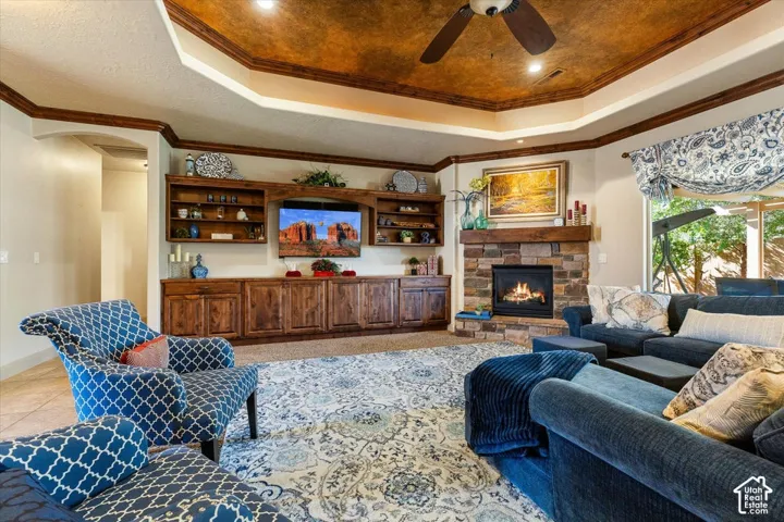 Tray ceiling with wood-finish trim, stone-clad fireplace, built-in wood cabinetry, integrated ceiling fan, and large windows