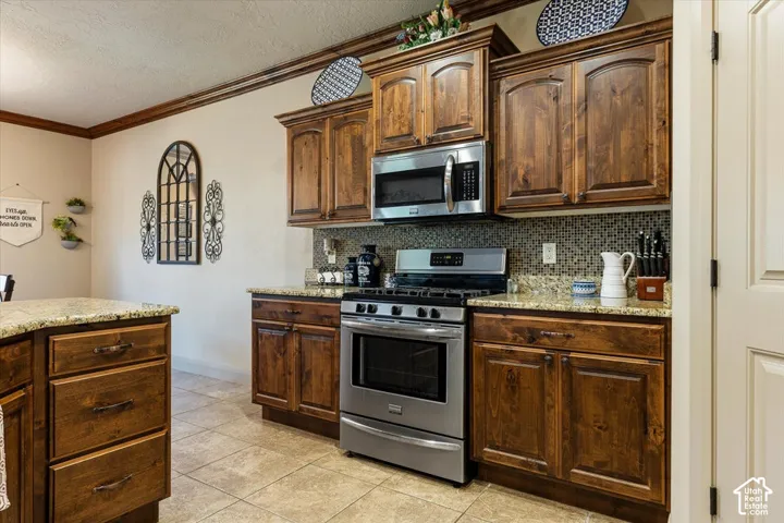 Brown wood-finish cabinetry with dark hardware, stainless steel appliances, granite countertops, mosaic tile backsplash, and light tile flooring