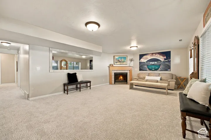 Carpeted living area featuring a fireplace with a wood mantel and tile surround, built-in wall mirror, and window with blinds