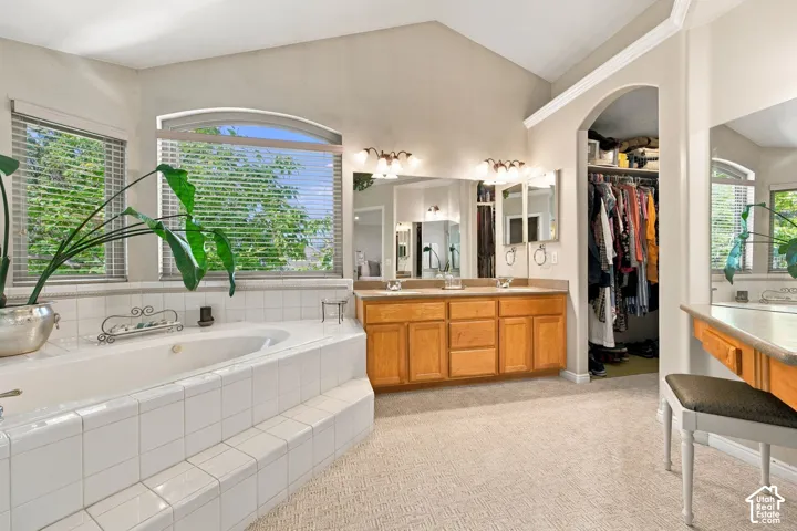 White tiled soaking tub, arched window, wood vanity with dual sinks, mirrored wall, and walk-in closet