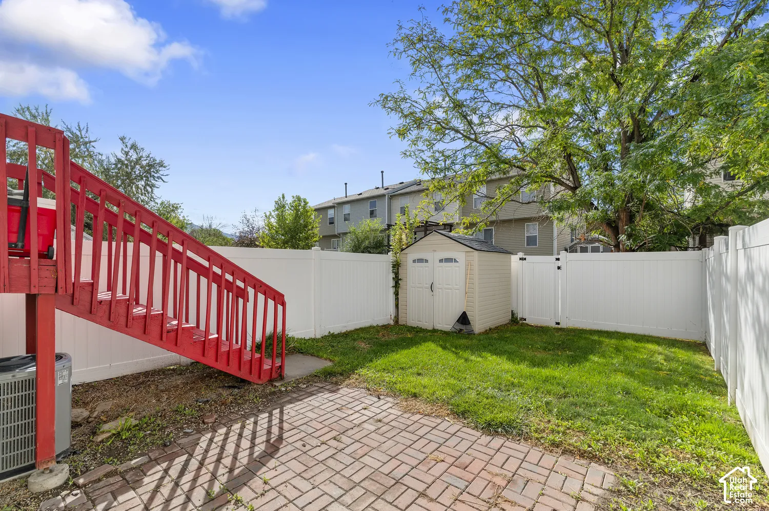 Fenced backyard featuring a red wood-finish staircase, brick paver patio, lush green lawn, and a storage shed