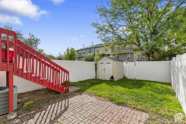 Fenced backyard featuring a red wood-finish staircase, brick paver patio, lush green lawn, and a storage shed