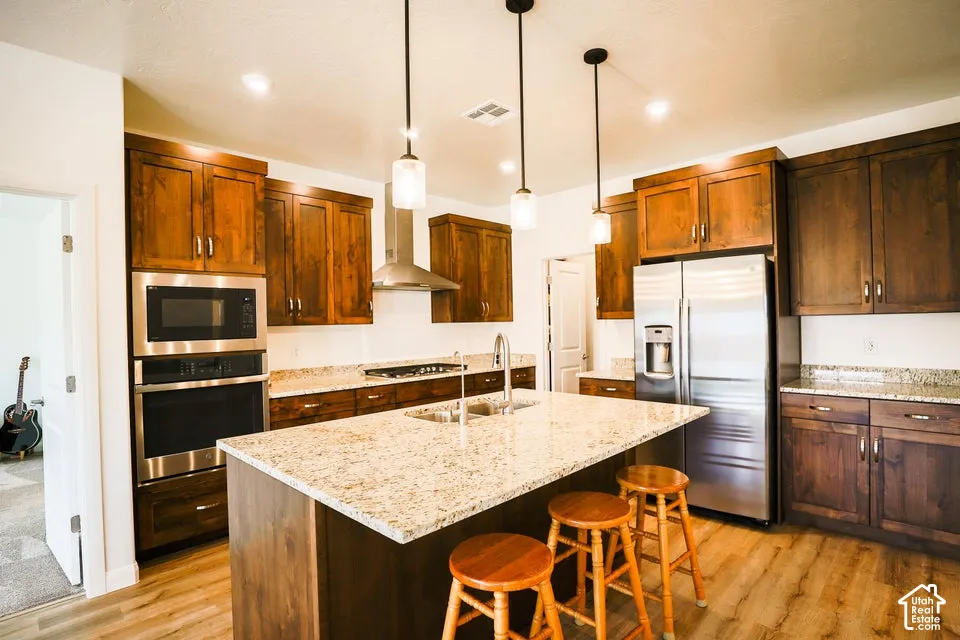 Kitchen featuring wood-finish flooring, stainless steel appliances, a large center island with a built-in sink, pendant lighting, and warm-toned cabinetry