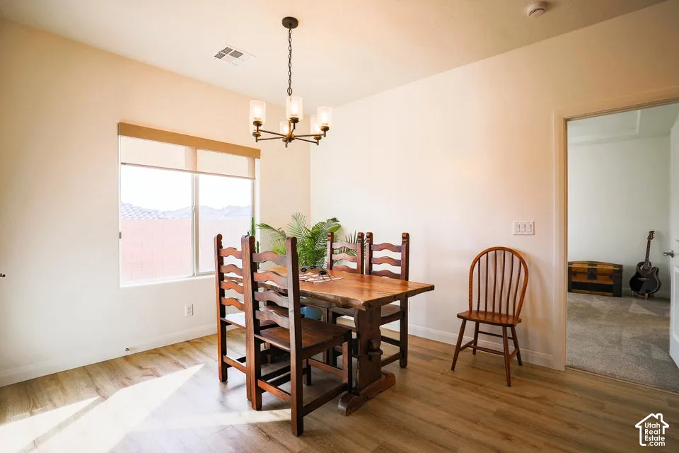 Light-toned walls with wood-finish flooring, featuring a window with roller shades, a ceiling-mounted chandelier, and an open doorway to a carpeted room