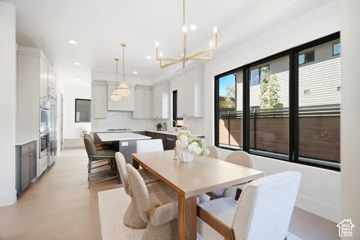 Open-concept kitchen and dining area featuring a wood-finish dining table, cream upholstered seating, light wood-finish flooring throughout, stainless steel appliances, white cabinetry, and large black-framed multi-panel windows and glass doors providing