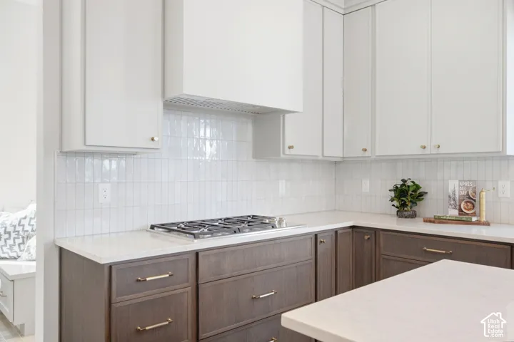 White upper cabinetry, wood-finish lower cabinetry, light-toned countertops, gas cooktop, and tile backsplash