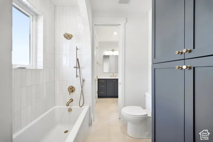 White tiled bath and shower combination with brass fixtures, a window above the tub, built-in storage cabinetry, and light-toned flooring