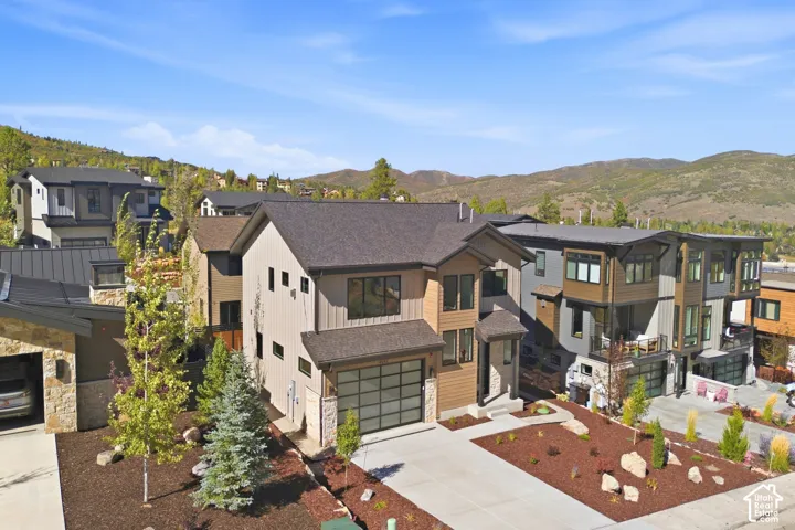 Modern exterior design featuring varied siding materials, attached two-car garage with a glass-paneled door, mature landscaping, concrete driveway, and expansive mountain scenery