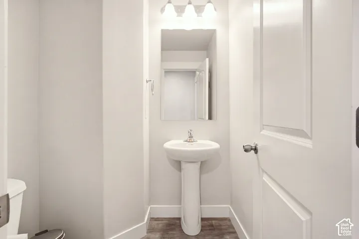 Pedestal sink with chrome faucet, mirror with overhead vanity lighting, white paneled door, and wood-finish flooring