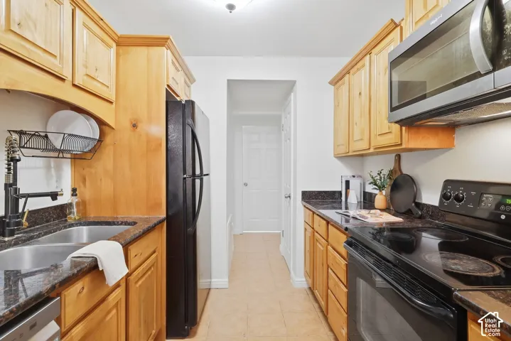 Galley kitchen featuring wood cabinetry, stone-finish countertops, stainless steel double basin sink, and black appliances