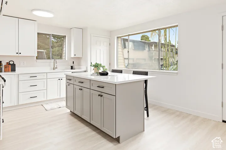 Modern kitchen with white cabinetry and dark hardware, featuring a central island with countertop seating, a stainless steel sink with a gooseneck faucet, light wood-finish flooring, and a large window