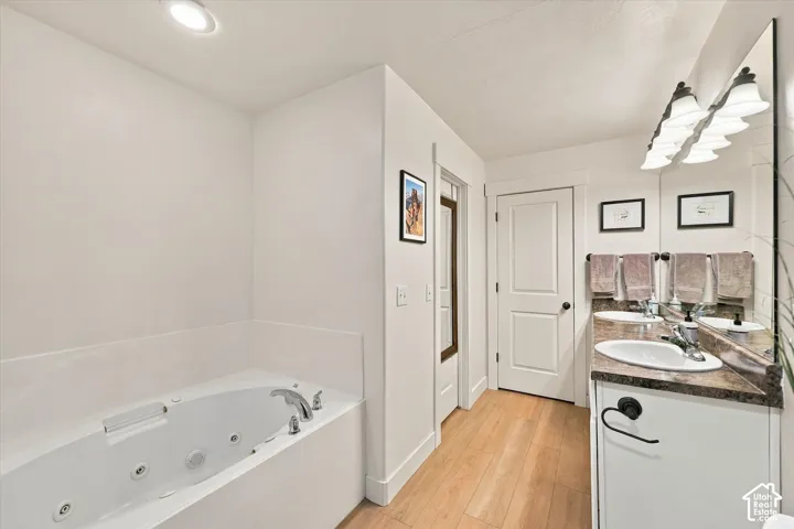 Soaking tub with chrome fixtures, dual vanity with undermount sinks, wood-finish flooring, and a large wall-mounted mirror