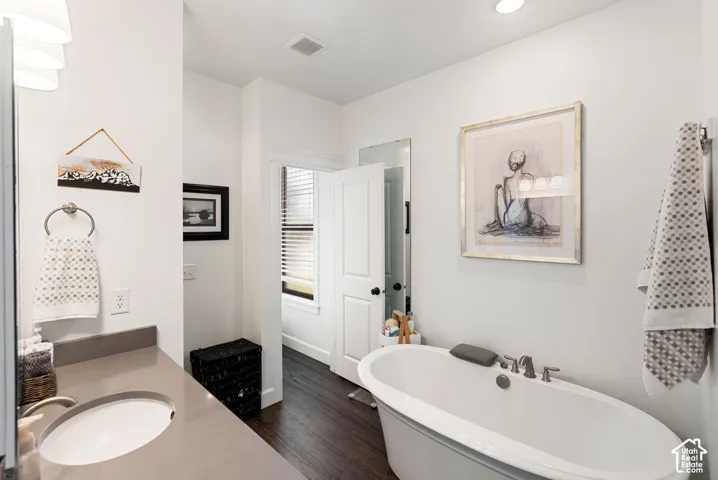 Freestanding soaking tub with brushed nickel fixtures, wood-finish flooring, recessed lighting, and a vanity with an integrated sink