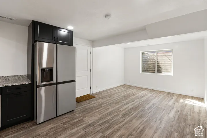 Stainless steel refrigerator, dark cabinetry, light-toned wood-finish flooring, white walls, and a window with a window well