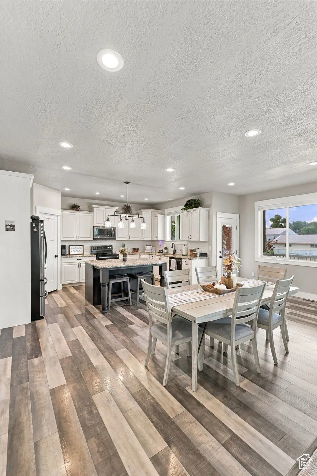 Open-concept kitchen and dining area featuring multi-tone wood-finish flooring, white cabinetry, recessed lighting, and a dark-toned kitchen island with seating