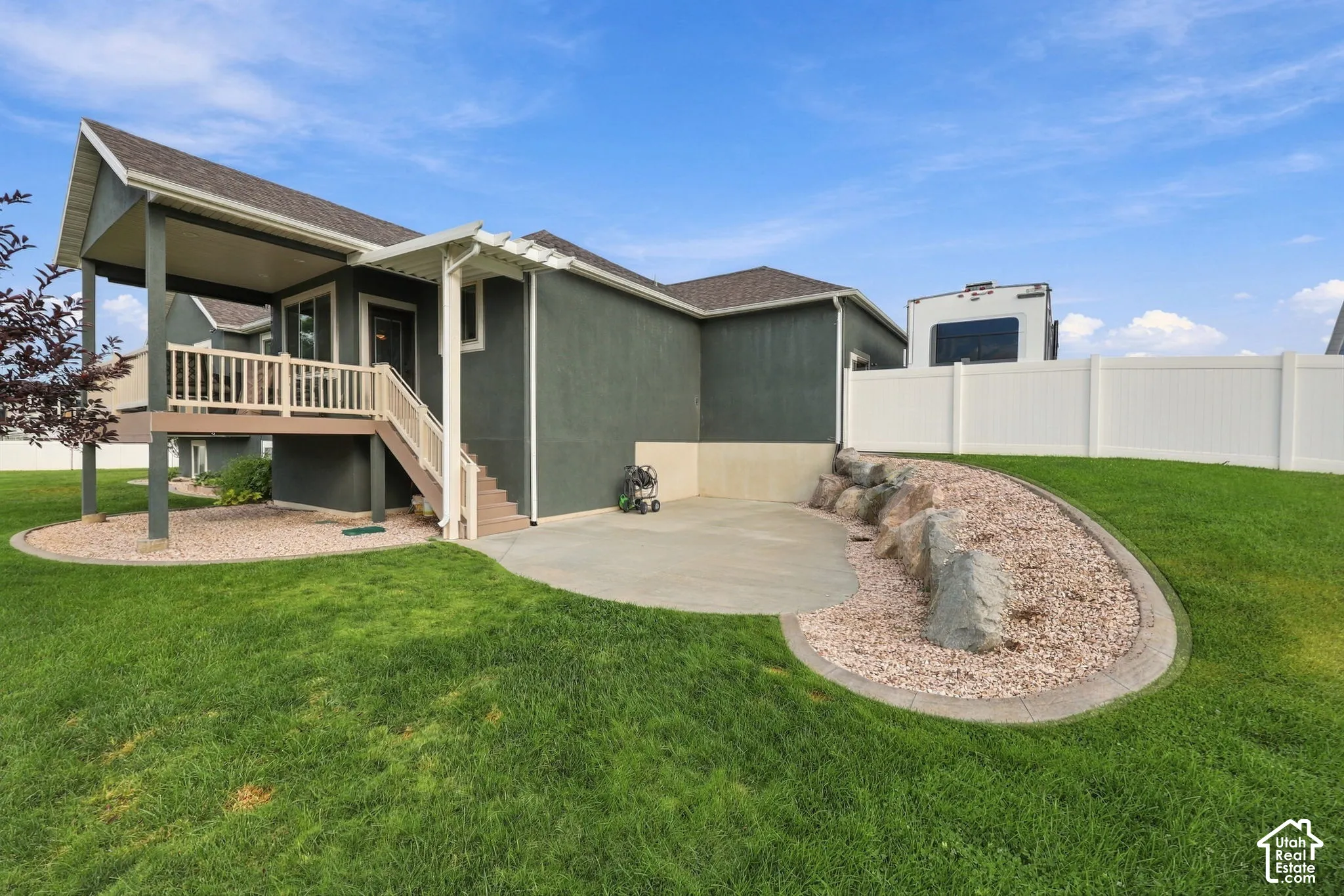 Expansive backyard featuring a covered deck, lower concrete patio, rock landscaping, and a white vinyl privacy fence