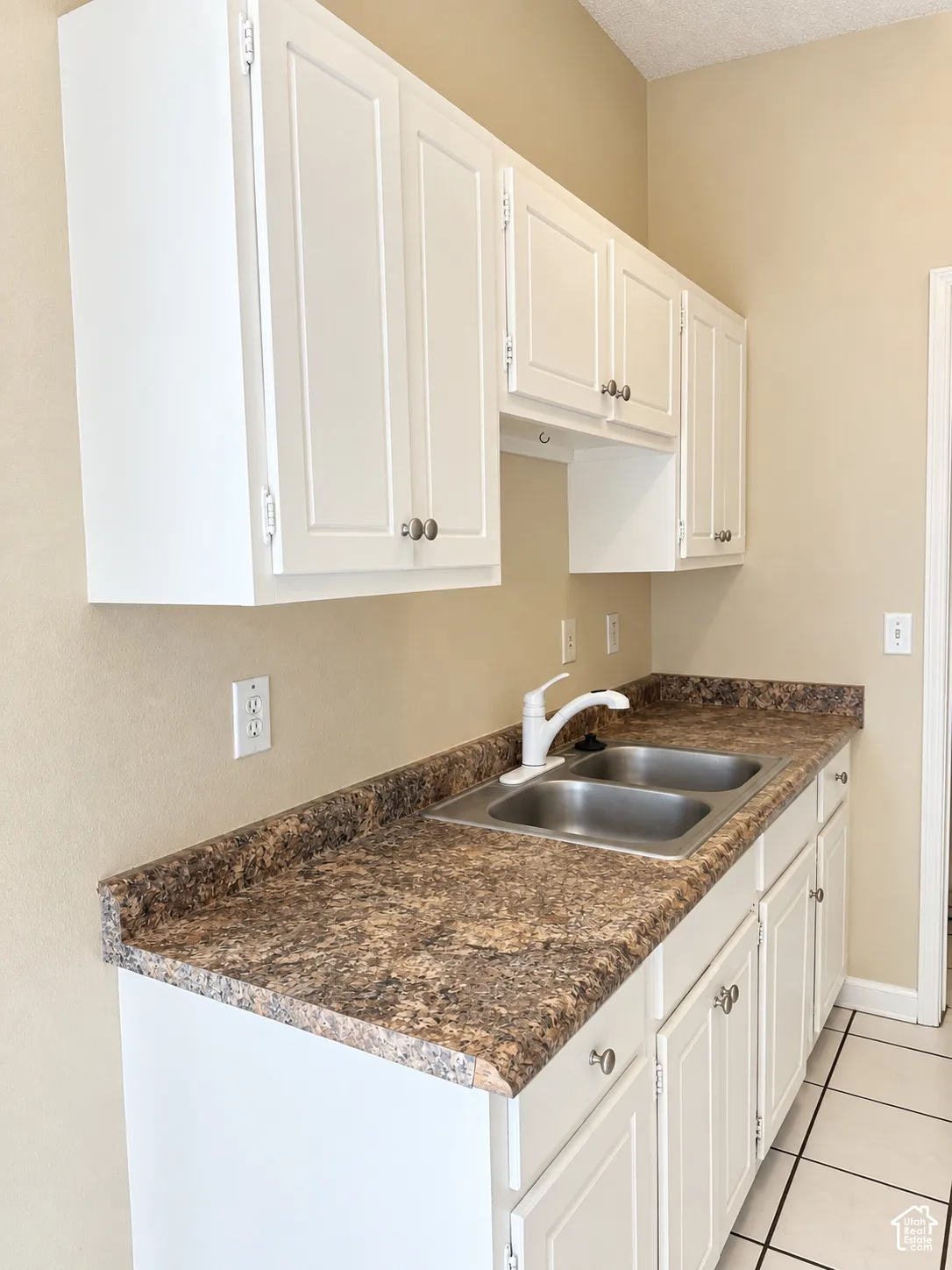 White shaker-style cabinetry with chrome hardware, dual-basin stainless steel sink, speckled laminate countertops, and tile flooring