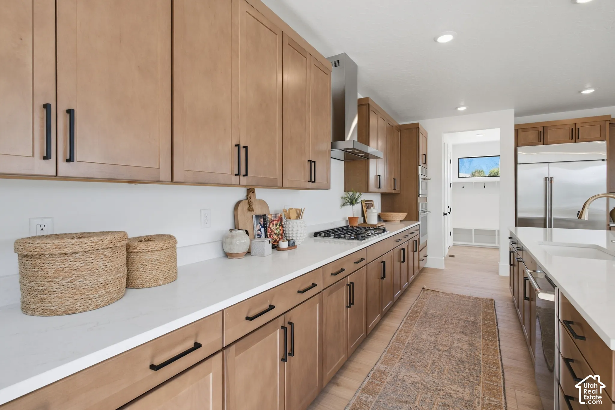 Light wood-finish cabinetry with matte black hardware, white countertops, built-in stainless steel appliances, recessed ceiling lights, and light wood-finish flooring