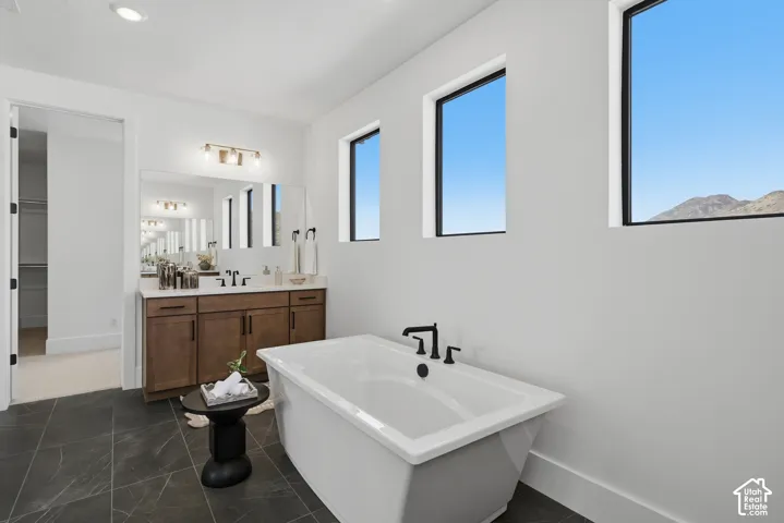 Freestanding soaking tub with a matte black faucet, dark tile flooring, a dual vanity with a light countertop, and multiple windows