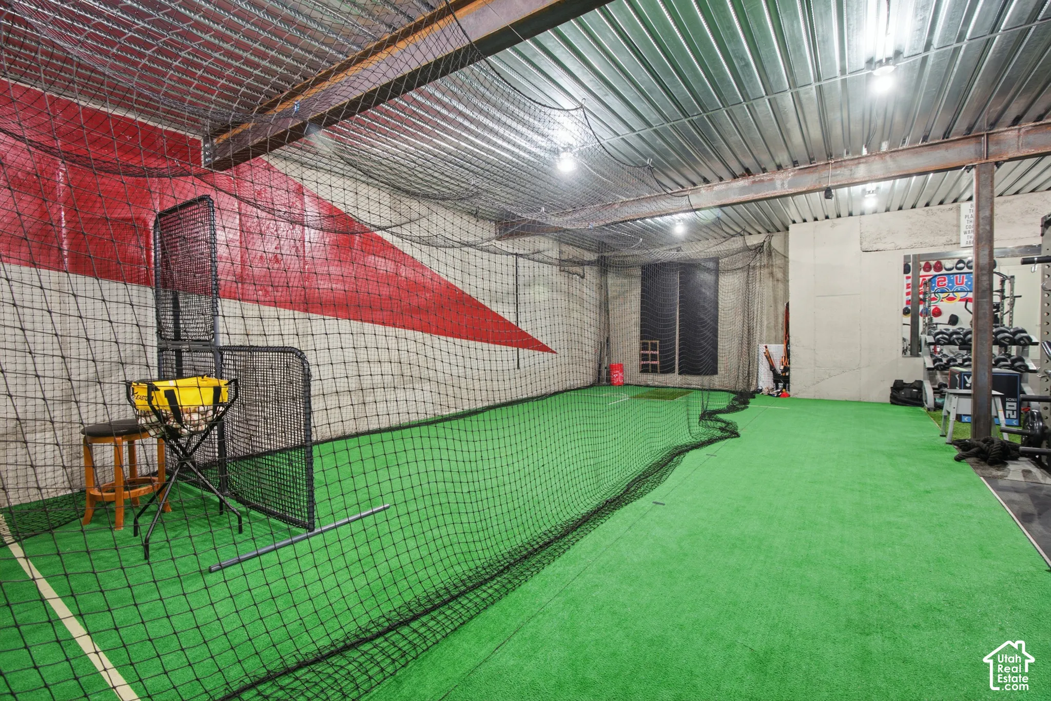 Indoor recreation space featuring artificial turf flooring, high ceilings with exposed metal beams, integrated lighting, and protective netting