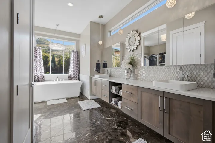 Freestanding soaking tub with a large window, dual vanity with vessel sinks, white countertops, and a tiled backsplash