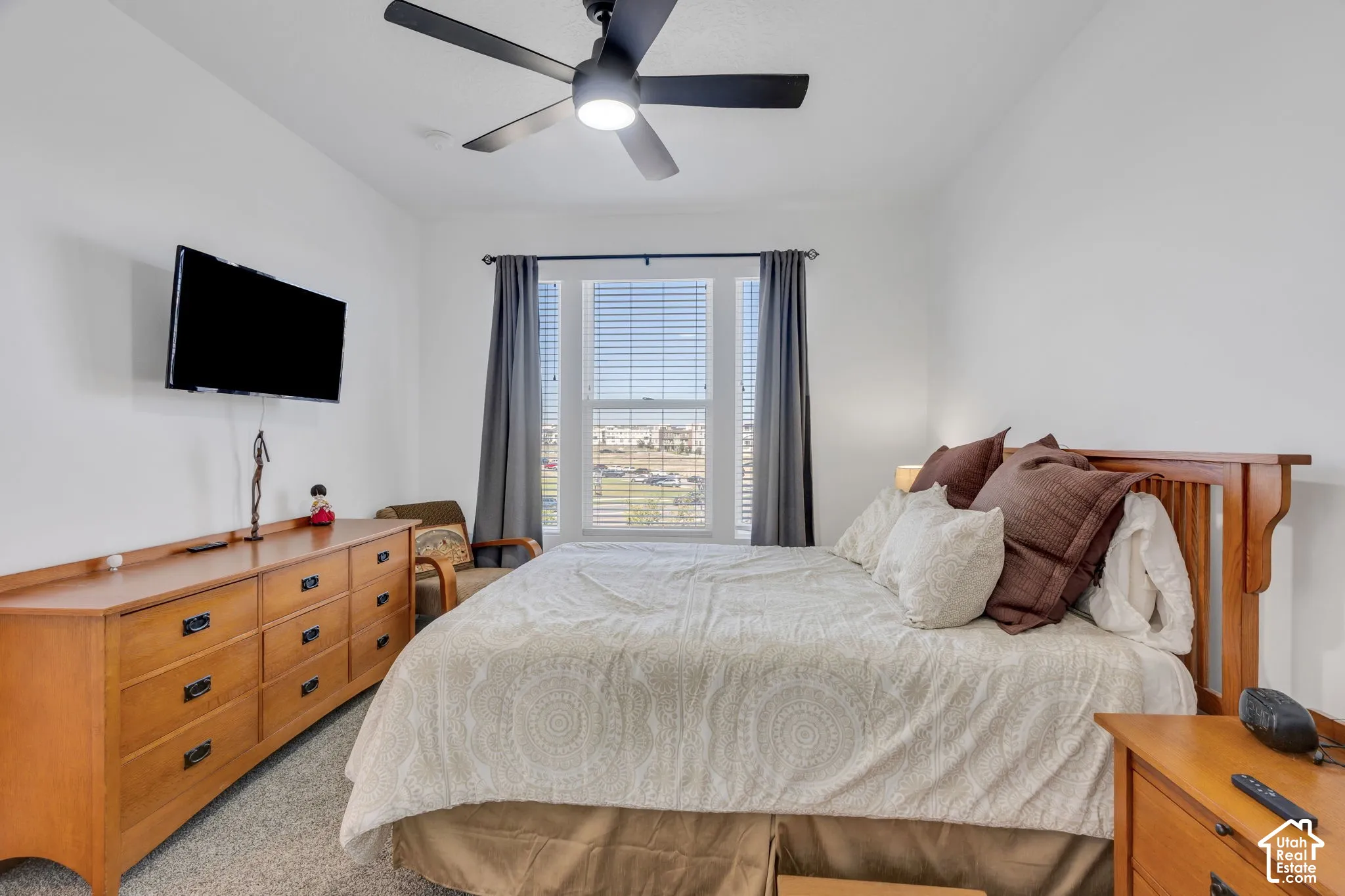 Carpeted bedroom featuring a ceiling fan, wood-finish dresser with dark hardware, and a window with blinds and drapes