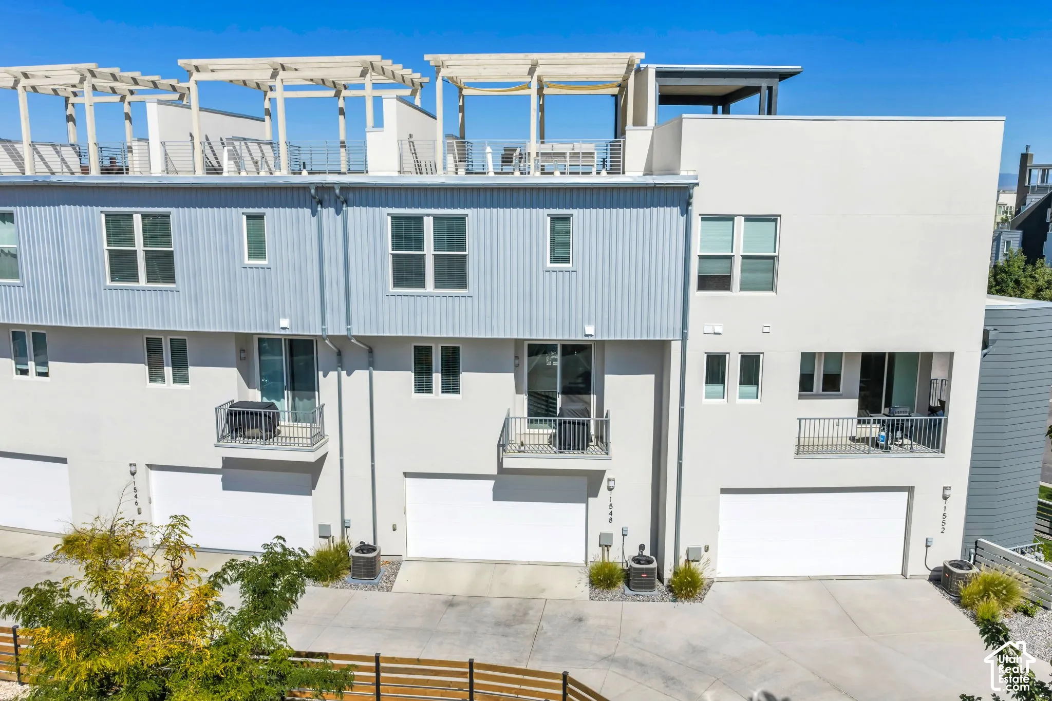 Modern townhouse exterior featuring multiple balconies with metal railings, a rooftop patio with pergolas, and integrated garage doors