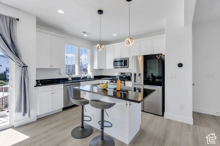 Modern kitchen featuring white cabinetry, black countertops, stainless steel appliances, a center island with seating, and light wood-finish flooring