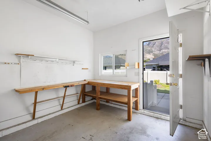 Concrete flooring, white painted walls, fluorescent lighting fixture, rear entry door, and a built-in workbench