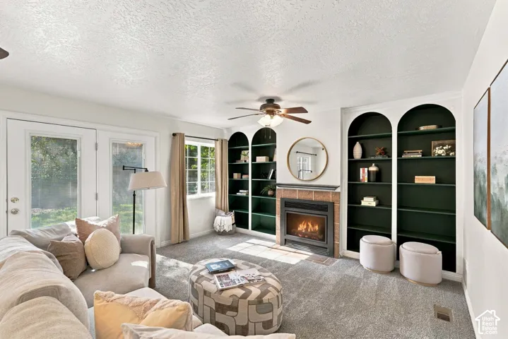 Recessed arched shelving units, tile-surround fireplace, dual-pane glass French doors, large window, and ceiling fan with integrated lighting