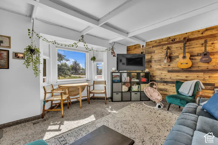 Exposed ceiling beams, bay window, wood-paneled accent wall, and carpeted flooring