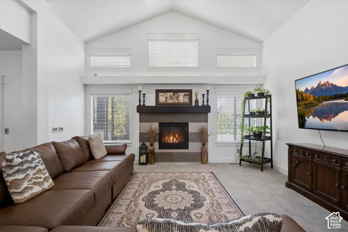 Vaulted ceiling, gas fireplace with tile surround, multiple windows, and light-toned carpet flooring