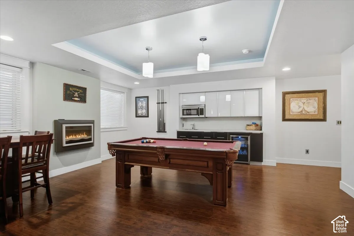 Wood-finish flooring throughout, featuring a recessed ceiling with accent lighting, a wall-mounted electric fireplace, and a wet bar with white upper cabinetry and a beverage refrigerator