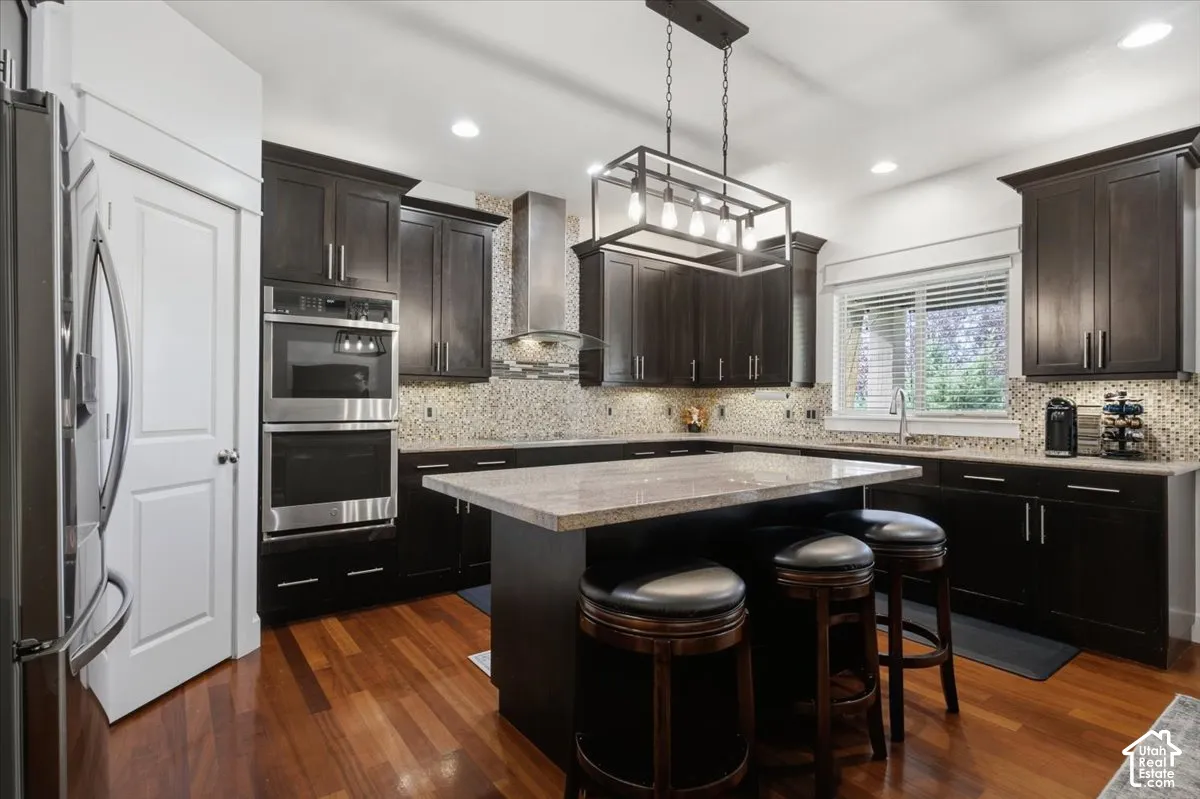 Hardwood flooring, dark wood cabinetry, stainless steel double ovens, stainless steel refrigerator, and a kitchen island with seating