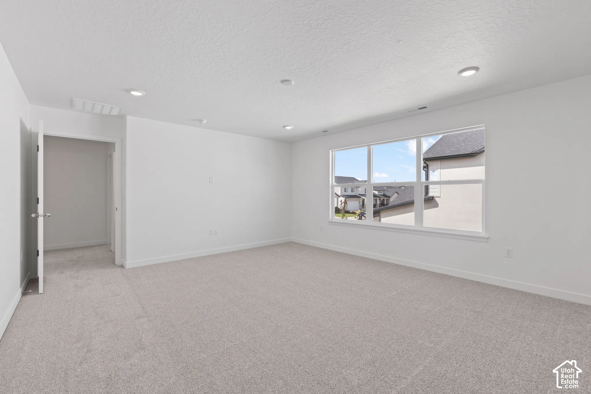 Spacious room with light-colored carpeting, multiple recessed ceiling lights, a large window, and white walls