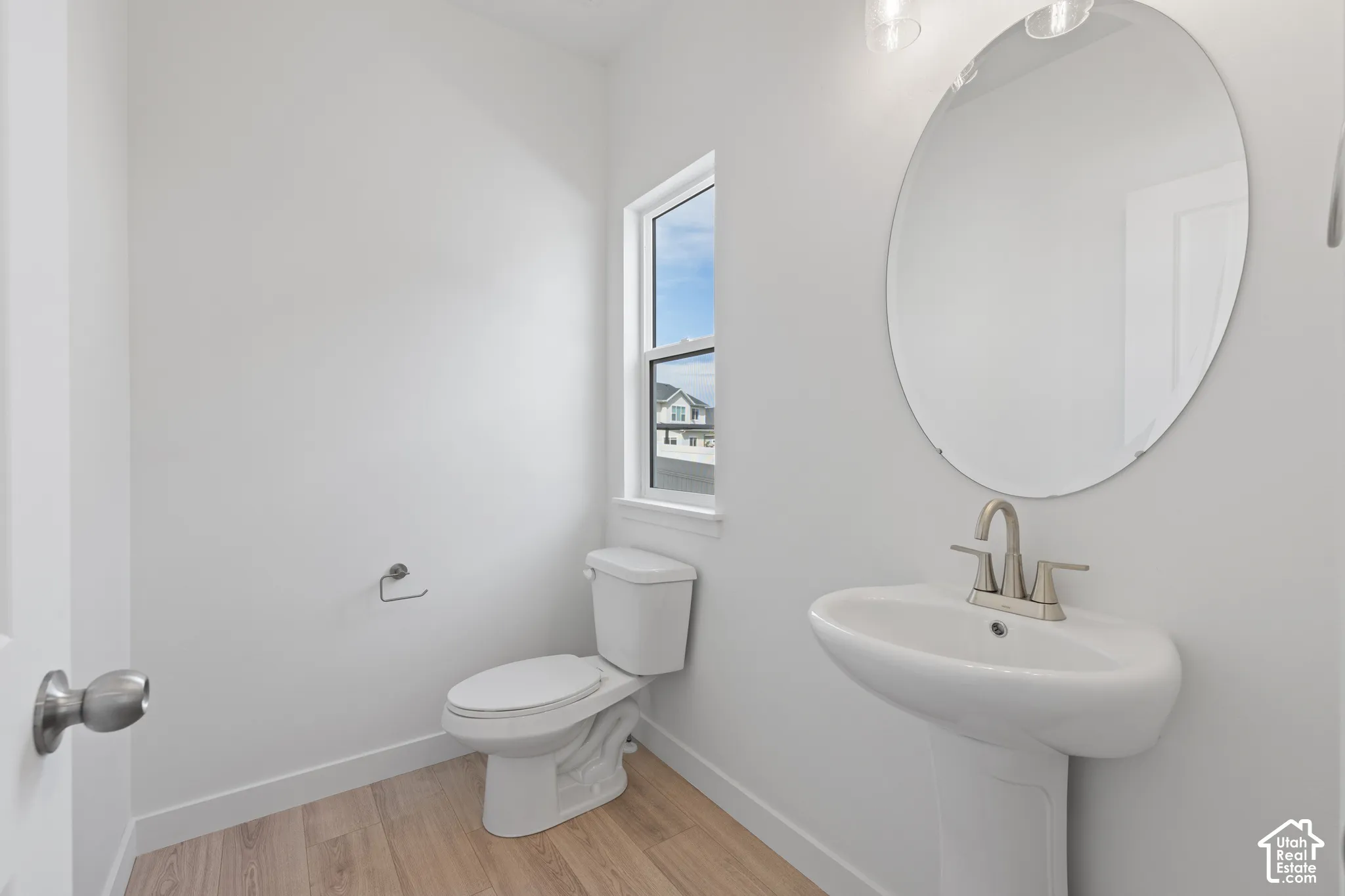 Pedestal sink with brushed nickel faucet, oval mirror, wall-mounted light fixture, and wood-finish flooring