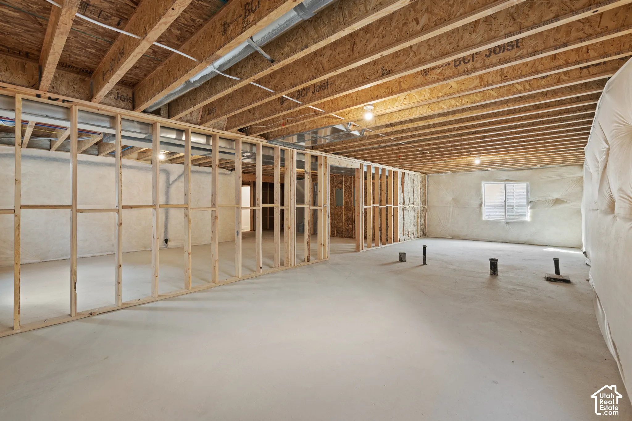 Unfinished basement with exposed ceiling joists, framed interior walls, concrete flooring, and a window with blinds