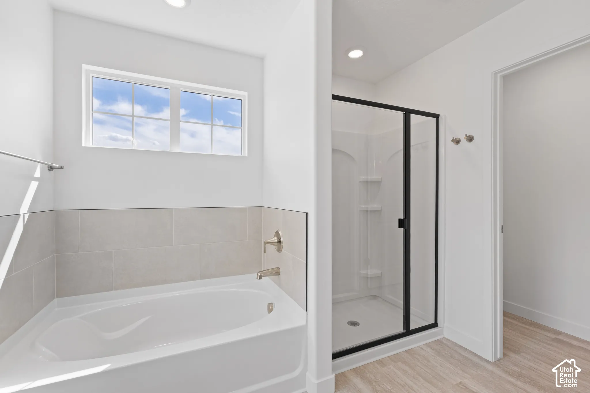 White soaking tub, light-toned wall tile, transom window, separate shower with black-framed glass enclosure, and wood-finish flooring